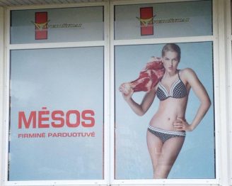 Ant mėsos gaminių parduotuvės kabanti reklama: moteris su bikiniu stovi seksualizuota poza ant peties užsimetusi didelį žalios mėsos gabalą. 