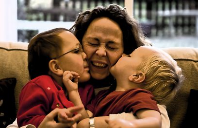 Mama apkabinusi du vaikus, kurie ją bučiuoja į skirtingus žandus