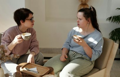 Mergina su Dauno sindromu kalbasi su kita mergina, abi merginos laiko po kavos puodelį, sėdi ant kėdžių